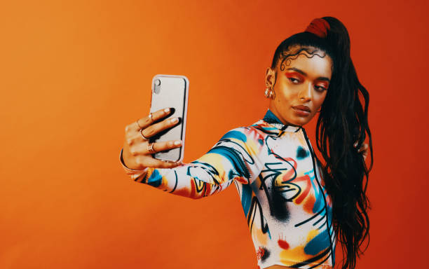 Studioaufnahme einer modischen Frau, die vor einem orangefarbenen Hintergrund ein Selfie macht