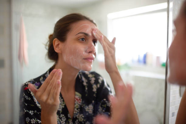 Femeie tânără în baie uitându-se în oglindă și având grijă de pielea feței.