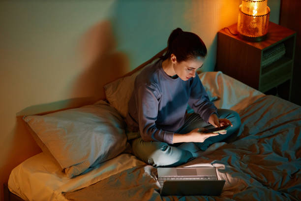 Korkeakulmainen muotokuva nuoresta naisesta, joka työskentelee myöhään kotona istuessaan sängyllä kannettavan tietokoneen ja älypuhelimen kanssa