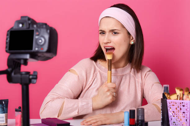 Indoor shot of charismatic artistic young blogger posing in front of camera around cosmetics, holding makeup tool in one hand, opening her mouth wide, looking directly at her camera. Shooting concept.