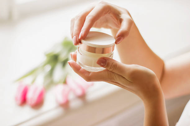 Closeup of young woman's hand holding jar of moisturizing cream in hands with spring flowers tulips on background. Gentle girl opening jar with face lotion in arms. Beauty treatment, skin or body care